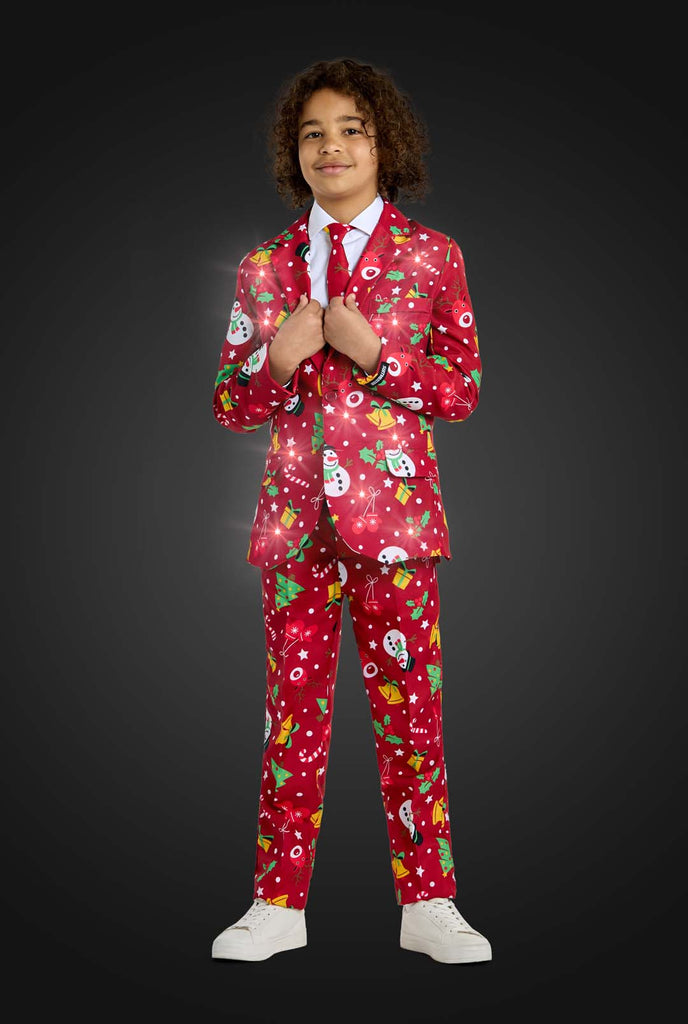 A child is wearing the Suitmeister "Christmas suit Red Icons Light Up," a festive red outfit adorned with snowmen, Christmas trees, stars, and presents. The child is smiling and light are on.