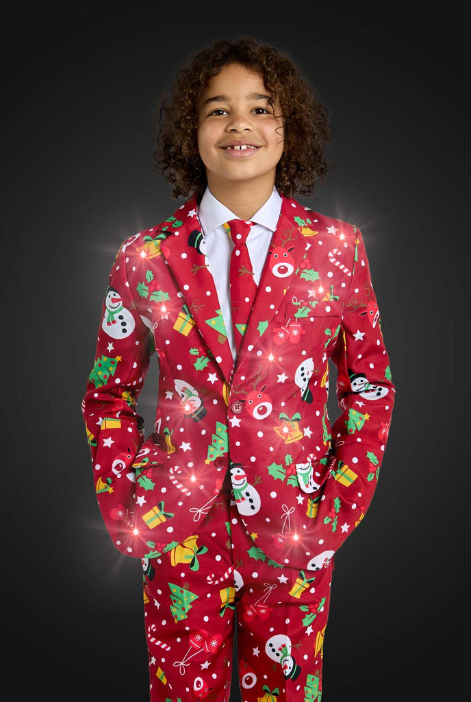 A child is wearing the Suitmeister "Christmas suit Red Icons Light Up," a festive red outfit adorned with snowmen, Christmas trees, stars, and presents. The child is smiling with their hands in pockets against a white background. Lights on jacket are on.