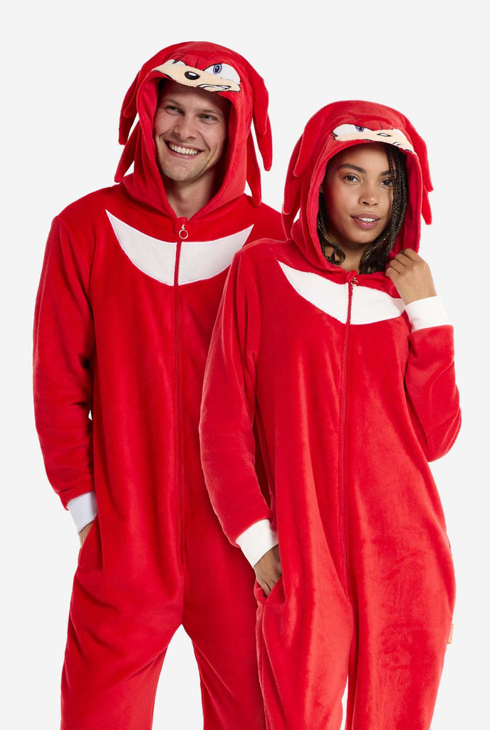 A man and woman are wearing a bright red Knuckles the Echidna-themed adult onesie from OppoSuits. The fun and cozy onesie features a zip-up front and an attached hood designed to resemble Knuckles' face, complete with expressive eyes and floppy dreadlocks. The suit is primarily red, with a white crescent pattern on the chest, reflecting Knuckles' iconic look from the Sonic the Hedgehog video game series. 