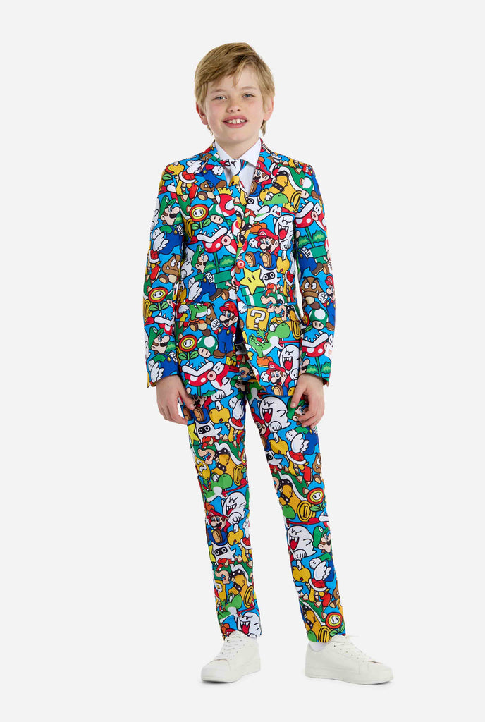 Image of teen wearing a Super Mario all-over print suit, consisting of blazer, pants and tie. Main color is blue, but there is also yellow, red and white. Full body image.