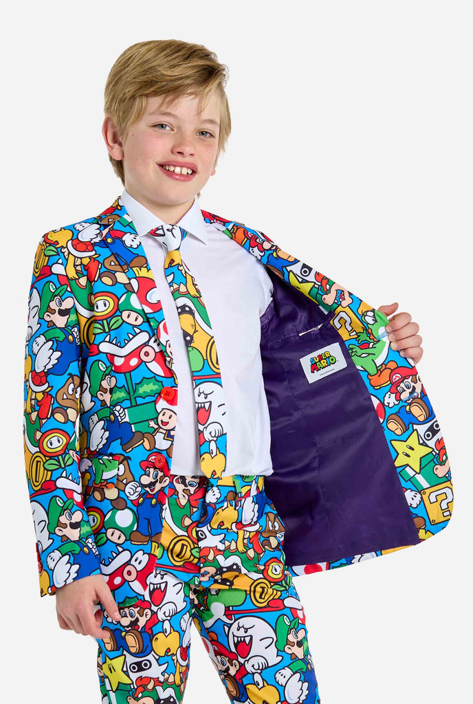 Image of teen wearing a Super Mario all-over print suit, consisting of blazer, pants and tie. Main color is blue, but there is also yellow, red and white. Holding ones side of the suit open.