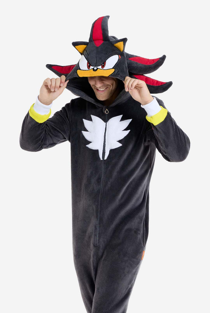 A man wearing a plush black onesie inspired by the popular hedgehog character Shadow, featuring spiky red and black accents on the hood, a white chest emblem, and yellow wrist details. The onesie is made from soft fleece material, offering comfort and a playful design for cosplay, lounging, or themed events. The person smiles while adjusting the hood, showcasing the character's recognizable face and bold design. Perfect for fans of gaming or animated characters.