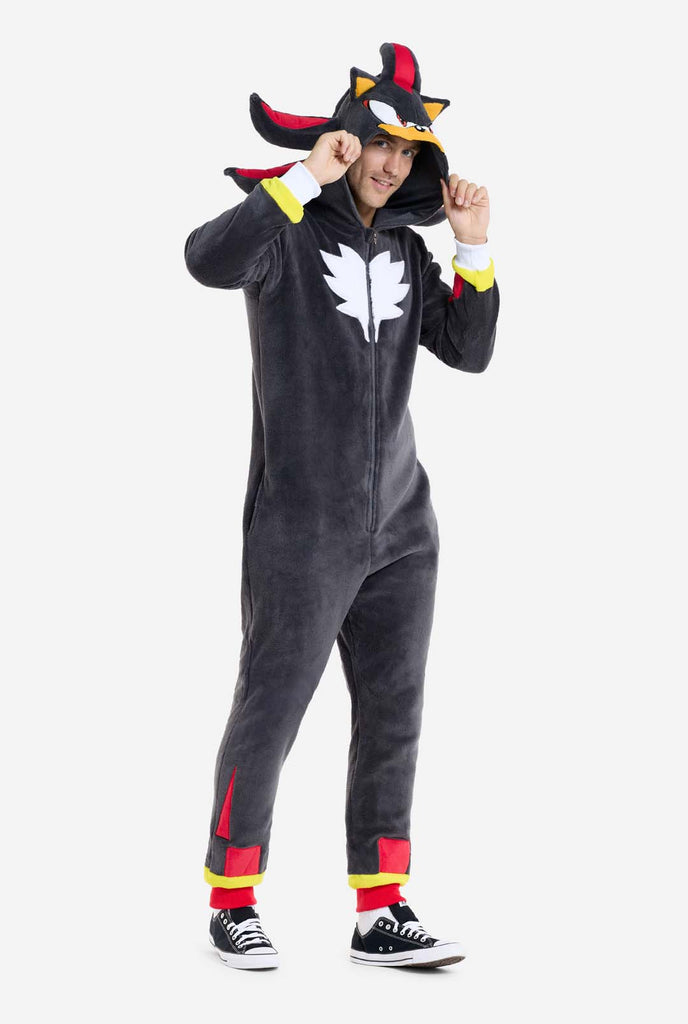 A man wearing a plush black onesie inspired by the popular hedgehog character Shadow, featuring spiky red and black accents on the hood, a white chest emblem, and yellow wrist details. The onesie is made from soft fleece material, offering comfort and a playful design for cosplay, lounging, or themed events. The person smiles while holding  the hood, showcasing the character's recognizable face and bold design. Perfect for fans of gaming or animated characters.