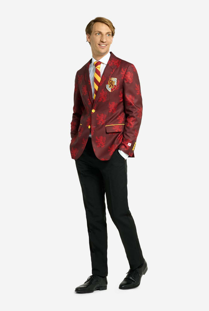 Image with light grey background and man wearing Harry Potter suit, Gryffindor styled. Suit consists of red blazer with Gryffindor logo and light red lions all over print, black pants, and red and yellow diagonal striped tie. Man holding hands in his pockets and looking side ways.