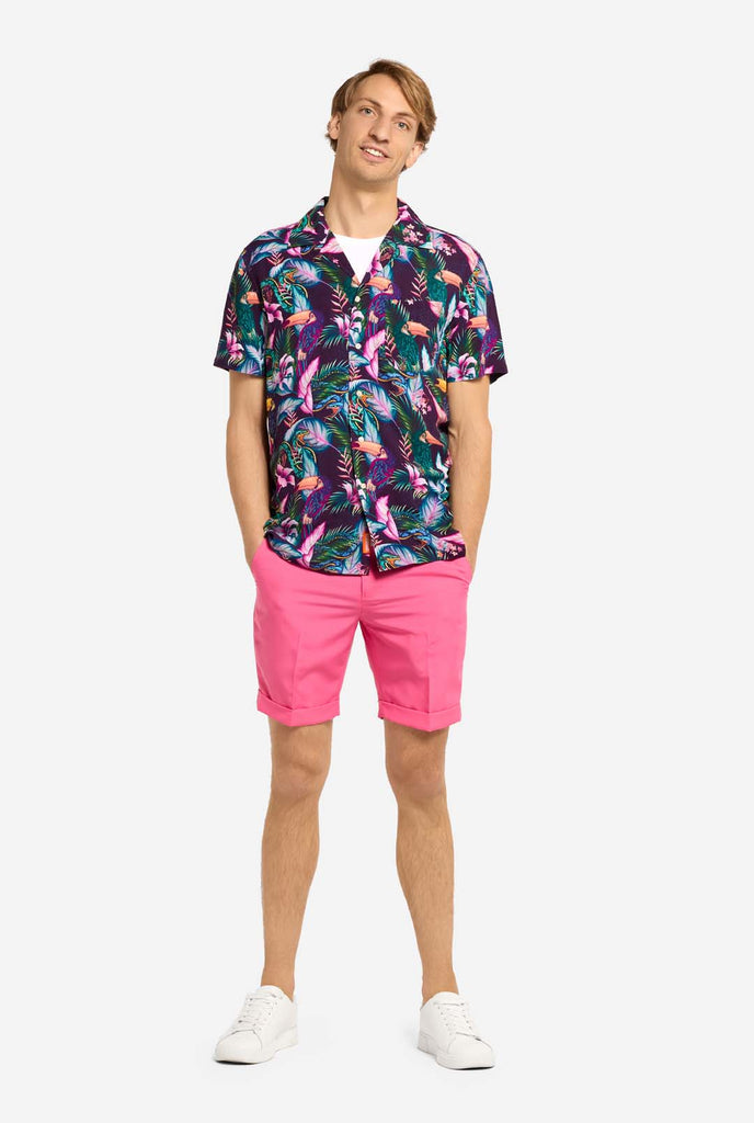 A man is wearing a purple Hawaiian shirt with a tropical print, resembling the jungle night live. Print consists of parrots, snakes, leaves and flowers. Also wearing pink shorts. He is smiling in the camera and holding holding hands in the pocket. Full body image.
