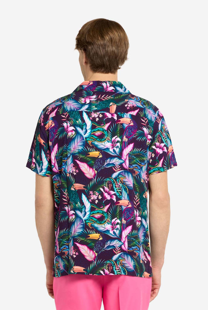A man is wearing a purple Hawaiian shirt with a tropical print, resembling the jungle night live. Print consists of parrots, snakes, leaves and flowers. Also wearing pink shorts. Showing the back of the shirt.