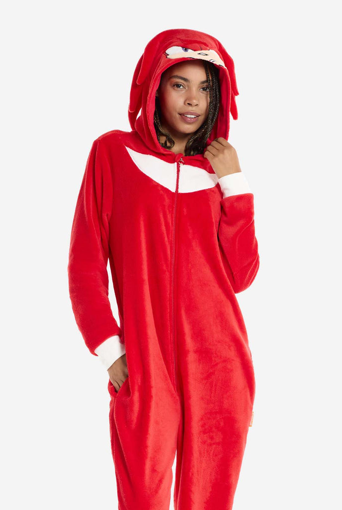 A woman wears a bright red Knuckles the Echidna adult onesie, a cozy and playful outfit inspired by the famous character from the Sonic the Hedgehog series. The onesie features Knuckles' signature white chest patch and includes a hood that showcases Knuckles' face, complete with his expressive eyes and long red dreads. The front zipper closure allows for easy wear, and the woman stands with one hand resting at her side while the other gently touches her hood. 