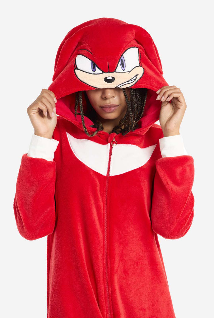 A woman wears a bright red Knuckles the Echidna adult onesie, a cozy and playful outfit inspired by the famous character from the Sonic the Hedgehog series. The onesie features Knuckles' signature white chest patch and includes a hood that showcases Knuckles' face, complete with his expressive eyes and long red dreads The front zipper closure ensures a comfortable fit, and the woman is holding the hood over her eyes The outfit, made from soft fleece material, is perfect for lounging or cosplay.