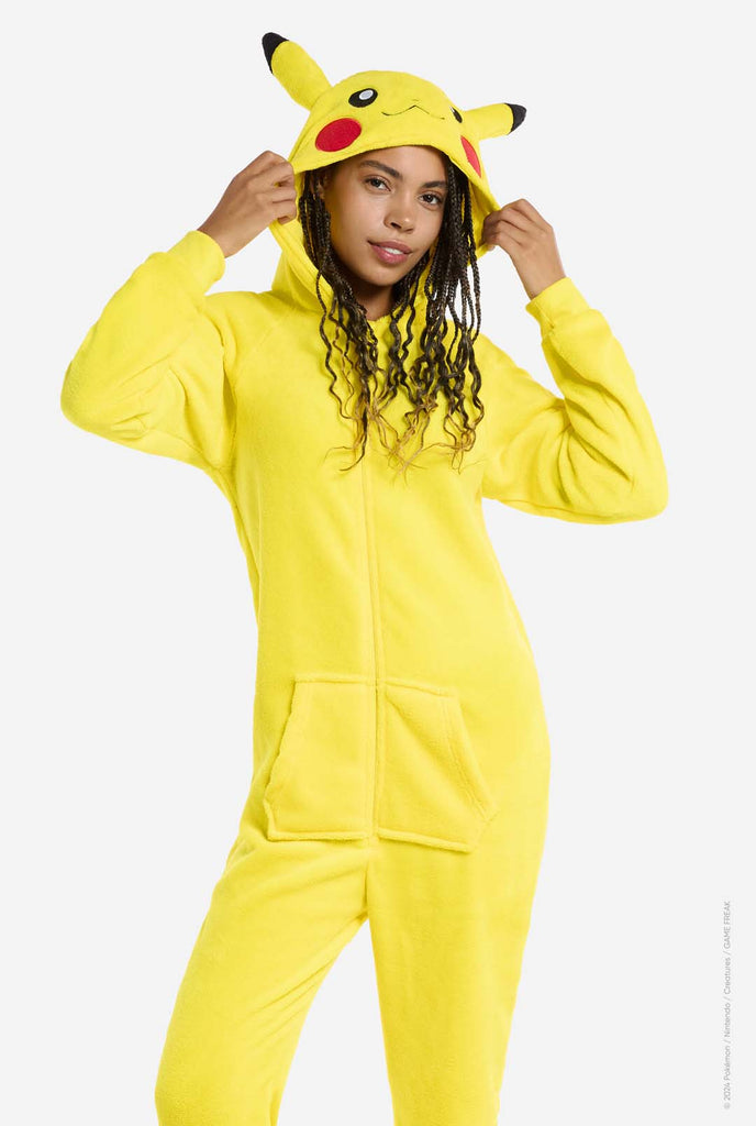 A woman is wearing a vibrant Pikachu onesie, designed to resemble the iconic Pokémon character. This unisex adult onesie is bright yellow with Pikachu’s recognizable face on the hood, featuring large black-tipped ears, black eyes, red cheeks, and a cute smile. The onesie has a convenient front zipper closure and two front pockets for functionality and comfort. Holding the hood with two hands. The soft, cozy material makes it perfect for lounging, cosplay events, or Pokémon-themed gatherings. 