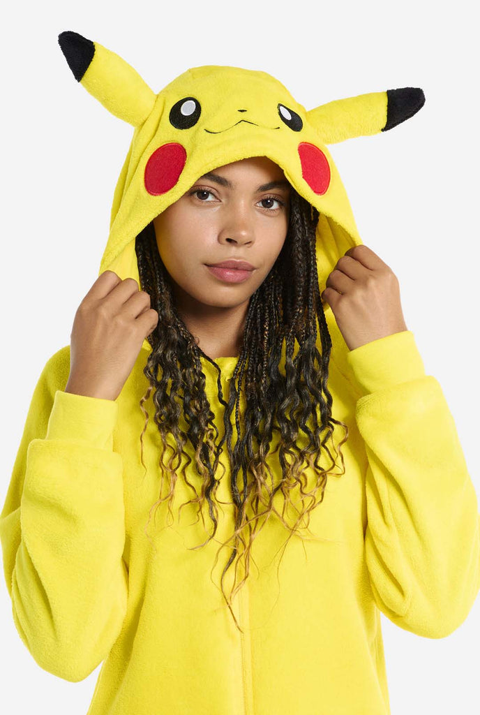 A woman is wearing a vibrant Pikachu onesie, designed to resemble the iconic Pokémon character. This unisex adult onesie is bright yellow with Pikachu’s recognizable face on the hood, featuring large black-tipped ears, black eyes, red cheeks, and a cute smile. The onesie has a convenient front zipper closure and two front pockets for functionality and comfort. Holding the hood with two hands. The soft, cozy material makes it perfect for lounging, cosplay events, or Pokémon-themed gatherings. Close up.