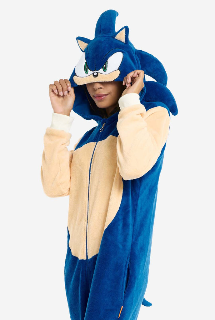 A woman is dressed in a cozy, blue Sonic the Hedgehog adult onesie, featuring soft beige patches on the front and sleeves to replicate Sonic's iconic look. The onesie has a hood designed with Sonic's recognizable face, including large green eyes, blue spikes, and pointy ears. The front zipper closure ensures a comfortable fit, and the woman is holding the hood over her eyes. Her expression is calm and relaxed. This Sonic the Hedgehog onesie is ideal for cosplay, lounging, or themed events. Side view.