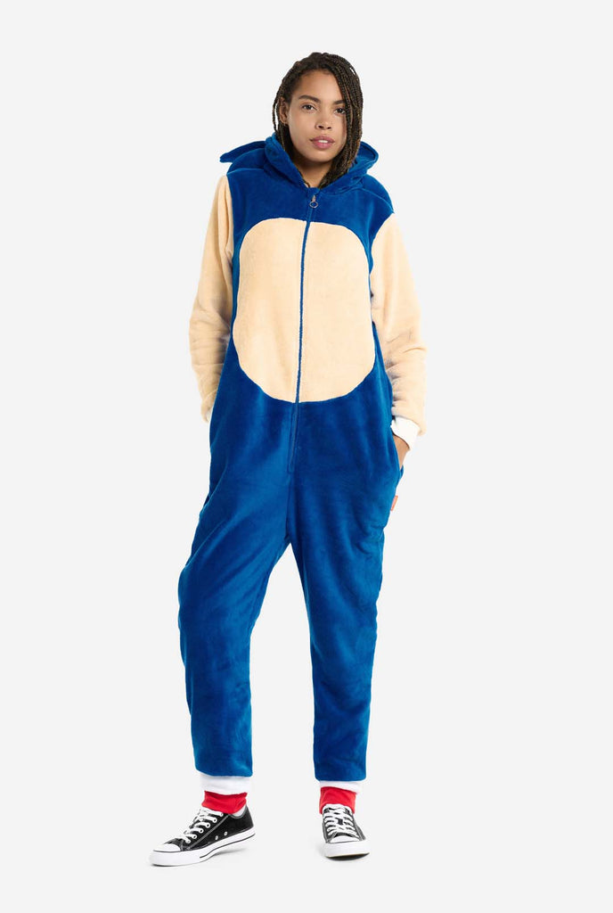 A woman is dressed in a cozy, blue Sonic the Hedgehog adult onesie, featuring soft beige patches on the front and sleeves to replicate Sonic's iconic look. The onesie has a hood designed with Sonic's recognizable face. The front zipper closure ensures a comfortable fit, and the woman stands with her hands in the onesie's side pockets. Her expression is calm and relaxed. This Sonic the Hedgehog onesie is ideal for cosplay, lounging, or themed events.