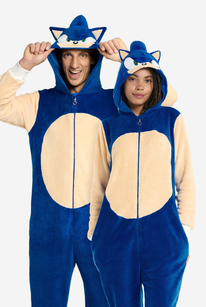 A man and woman wearing a playful blue and beige Sonic the Hedgehog-themed adult onesie from OppoSuits. The cozy onesie features a zip-up front and an attached hood designed to resemble Sonic’s iconic face, complete with ears and an expressive gaze. The blue body of the suit contrasts with a beige belly, mimicking Sonic's classic look. 