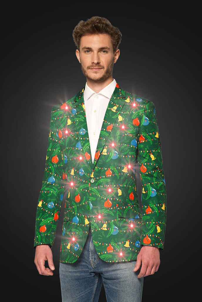 A man dressed in the Suitmeister Christmas Green Tree Jacket Light Up, featuring vibrant Christmas lights and patterns, stands against a dark gradient background. He complements the blazer with a white shirt, blue jeans, and black sneakers.