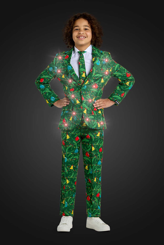 A child confidently stands with their hands on their hips, smiling against a dark background while wearing the Suitmeister Christmas suit Green Tree Light Up, a festive green outfit featuring vibrant Christmas lights and patterns like red ornaments and yellow stars.