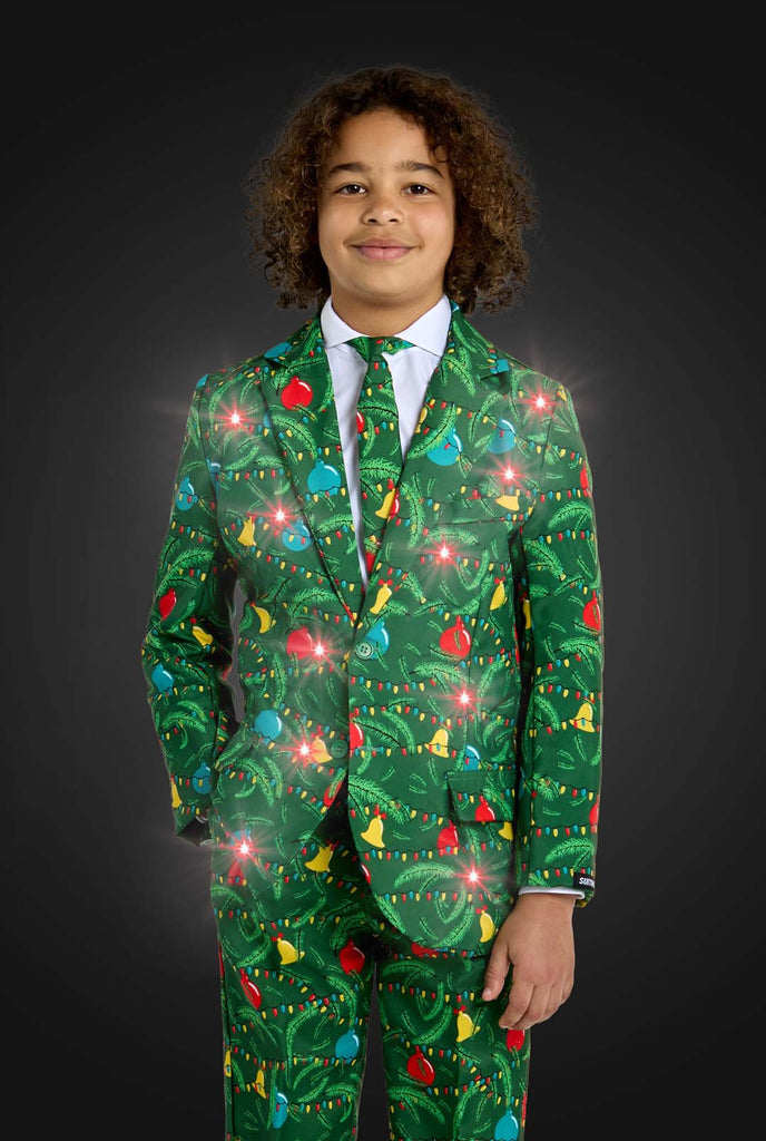 A child confidently stands with their hands on their hips, smiling against a dark background while wearing the Suitmeister Christmas suit Green Tree Light Up, a festive green outfit featuring vibrant Christmas lights and patterns like red ornaments and yellow stars.