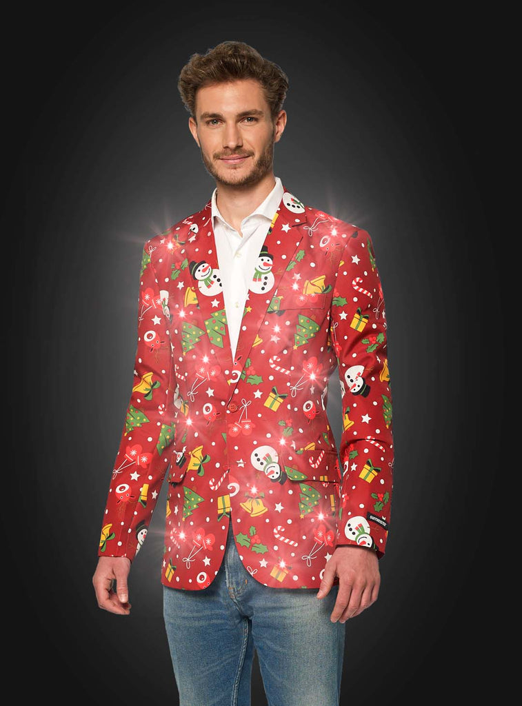 A person stands wearing the Christmas Red Icons Jacket Light Up by Suitmeister, featuring festive designs like snowmen and snowflakes. They pair it with blue jeans and black sneakers, smiling against a dark background as the jacket illuminates.