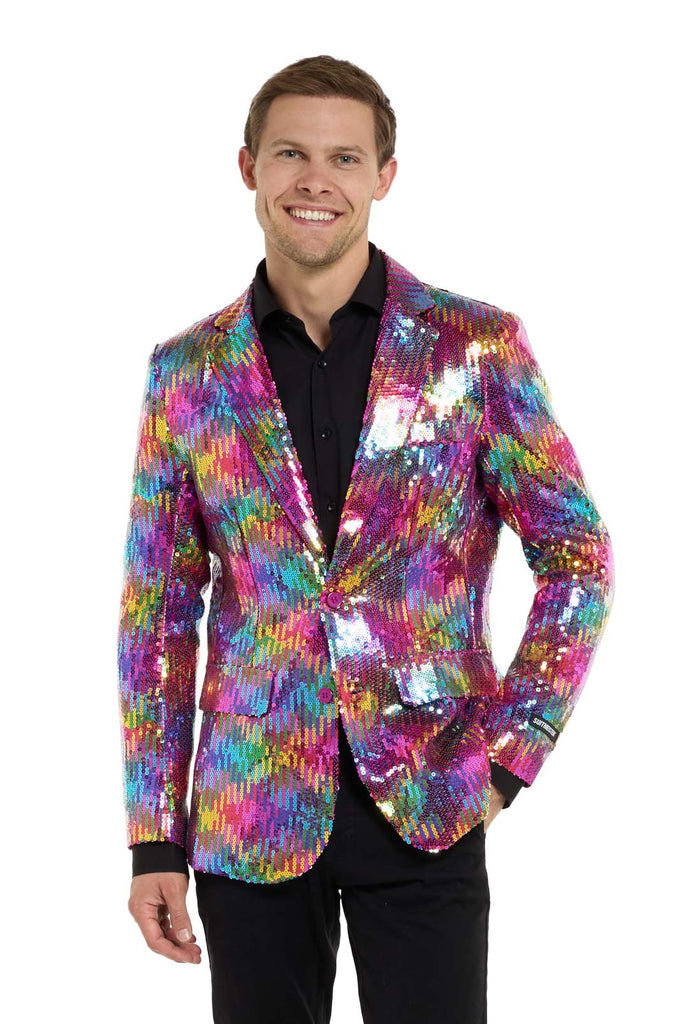 Image with white background and man wearing rainbow colored sequins blazer. Man also wearing black shirt and black pants. One hand in his pocket and  looking in the camera.