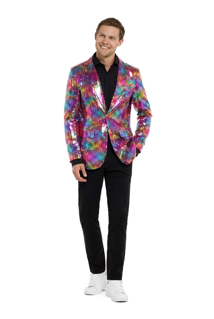 Image with white background and man wearing rainbow colored sequins blazer. Man also wearing black shirt and black pants. One hand in his pocket and  looking away from the camera. Full body image.