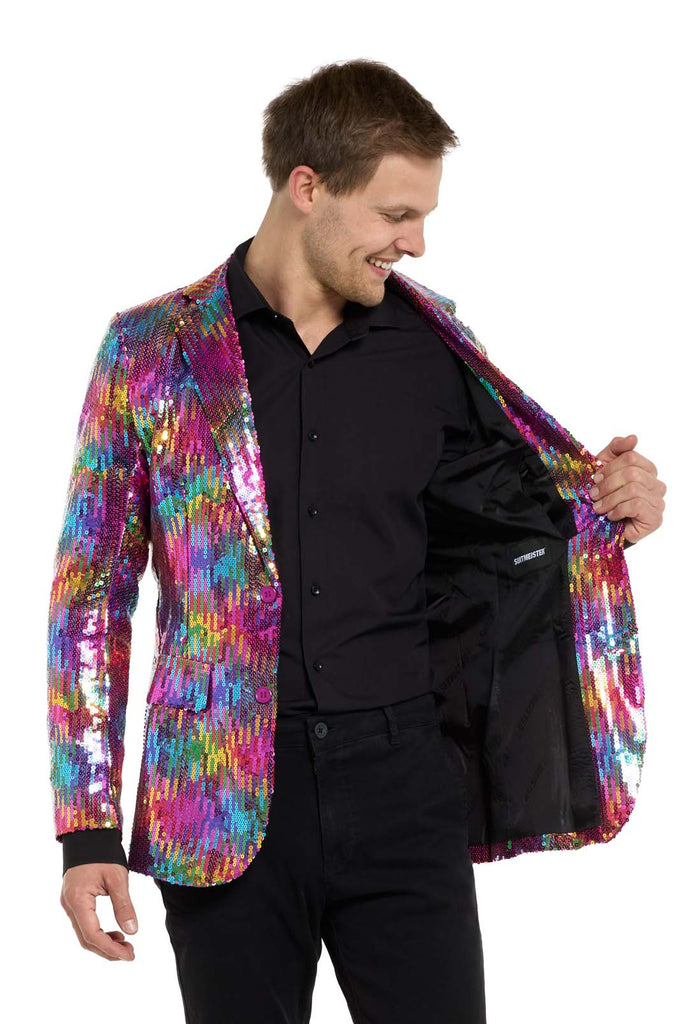 Image with white background and man wearing rainbow colored sequins blazer. Man also wearing black shirt and black pants. Holding one side of the jacket open.