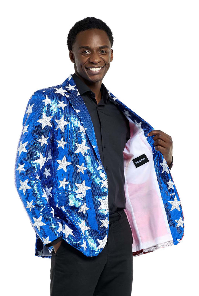 Image with white background and man wearing USA flag-colored red, blue and white sequins blazer. The man is also wearing black shirt and black pants. Holding one side of the blazer open and smiling in the camera.