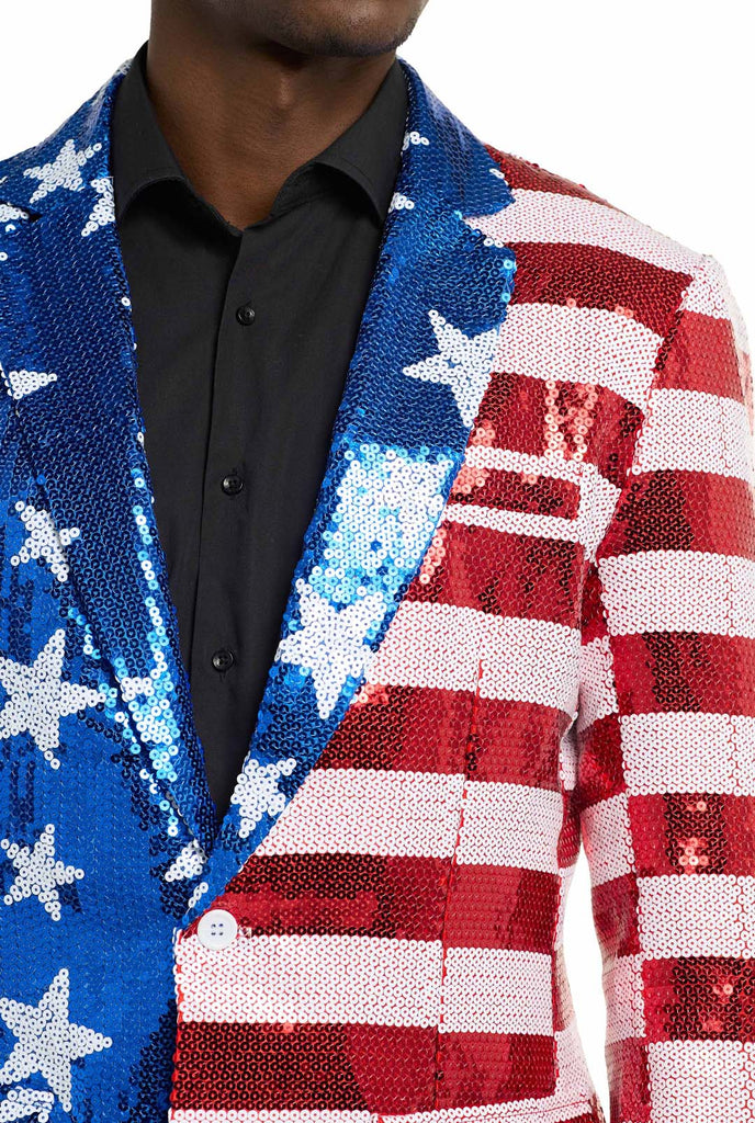 Image with white background and man wearing USA flag-colored red, blue and white sequins blazer. The man is also wearing black shirt and black pants. Chest close up.