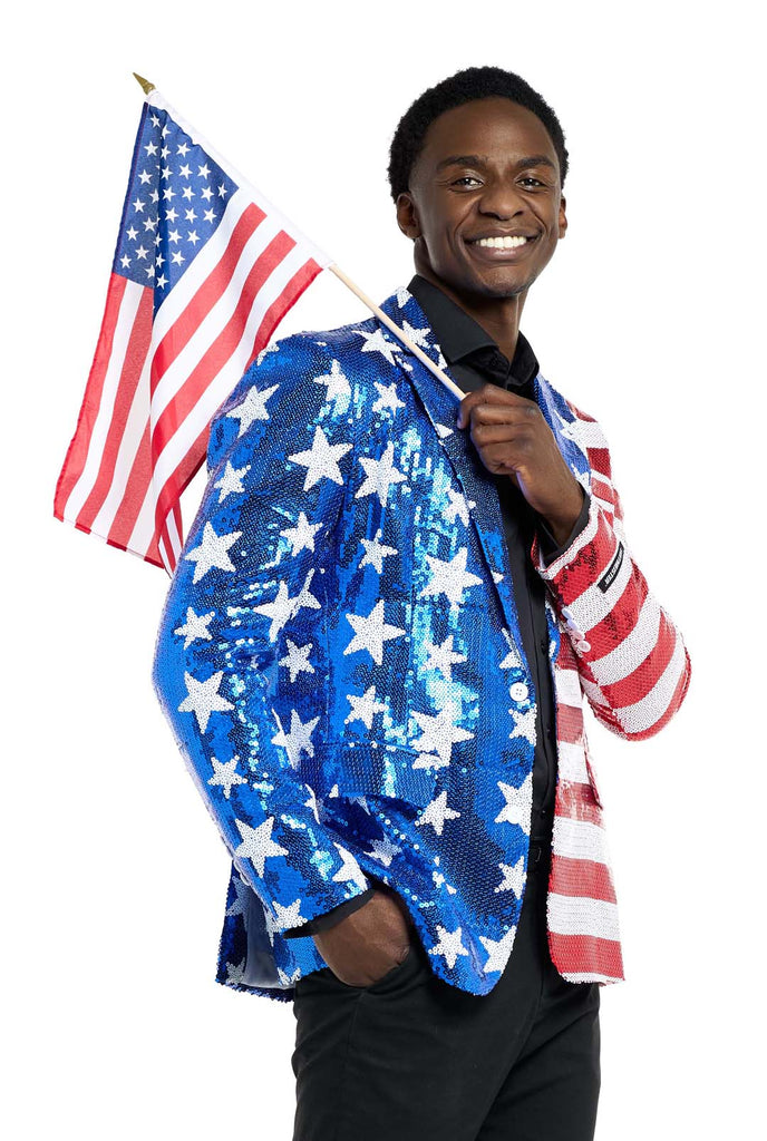 Image with white background and man wearing USA flag-colored red, blue and white sequins blazer. The man is also wearing black shirt and black pants. The man is holding a small USA flag over his shoulder.