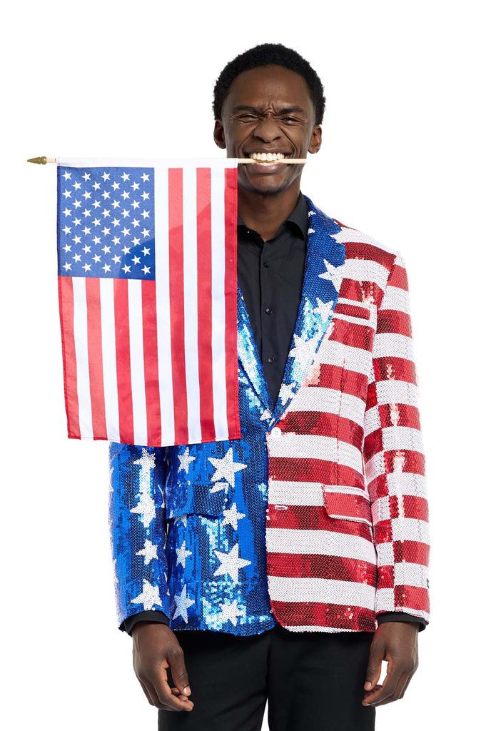 Image a white background and man wearing a USA flag-colored red, blue, and white sequins blazer. The man is also wearing a black shirt and black pants. The man is holding a small USA flag between his teeth.